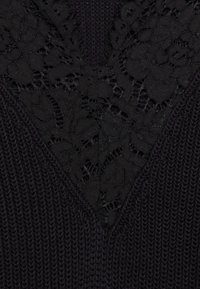 Giacchetto in maglia nera con scollo a V e inserto in pizzo nero. Il materiale lavorato a maglia presenta un contrasto con il dettaglio in pizzo liscio.