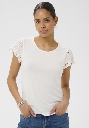 Femme aux cheveux bruns tirés en arrière, portant une chemise blanche à manches en dentelle, un jean bleu, des boucles d'oreilles argentées en forme d'anneau et un bracelet argenté, mains dans les poches.