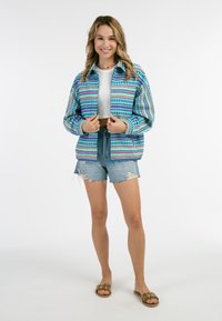 Giacca colorata a fantasia blu, verde e rosa, indossata sopra una maglietta corta bianca, con pantaloni shorts in denim strappati e sandali decorati con fiori.