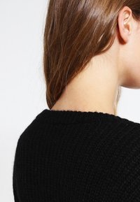 Hnědé vlasy schované za uchem, oblékající černý pletený svetr, ukazující stranu a zadní část krku a hlavy na jednobarevném pozadí.