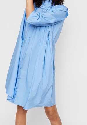 Kobieta ubrana w luźną, jasnoniebieską sukienkę na guziki z rękawami do połowy przedramion, stojąca na jednolitym białym tle.