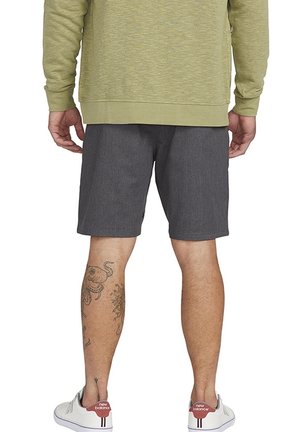 Uomo che indossa pantaloni corti grigi, felpa verde oliva, sneakers bianche New Balance, e tatuaggi sulle gambe visibili sul polpaccio destro visti da dietro.
