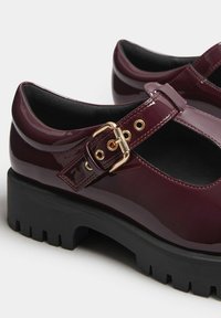 Zapatos Mary Jane de charol burdeos con suela de plataforma negra gruesa, que cuentan con una hebilla dorada y orificios de ventilación cerca de la correa.