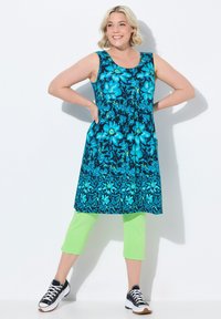 Květinové tunikové šaty z modré a černé látky s kapsami, spárované s jasně zelenými zkrácenými kalhotami a černými teniskami.