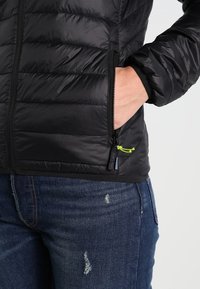 Svart puffjacka med kviltat mönster, dragkedjeficka med en grön dragrem, bärs över slitna blå jeans.