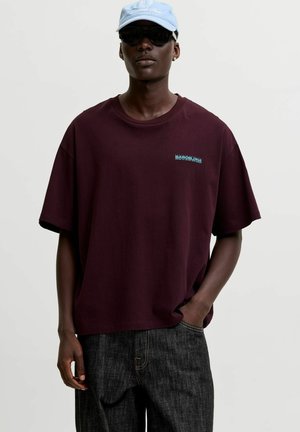 Mand iført mørkelilla overdimensioneret t-shirt, mørke jeans, lyseblå kasket, sorte solbriller og med den ene hånd i lommen mod ensfarvet baggrund.