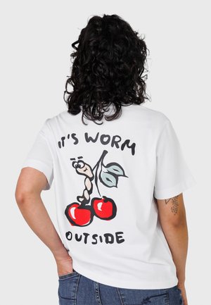 Osoba o kręconych ciemnych włosach, ubrana w białą koszulkę z kreskówkowym robakiem na dwóch czerwonych wiśniach i napisem „It's worm outside” na plecach.