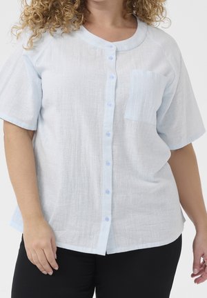 Krullend persoon die een lichtblauw, kort mouwig, overhemd met borstzak en een zwarte broek draagt tegen een egale achtergrond.