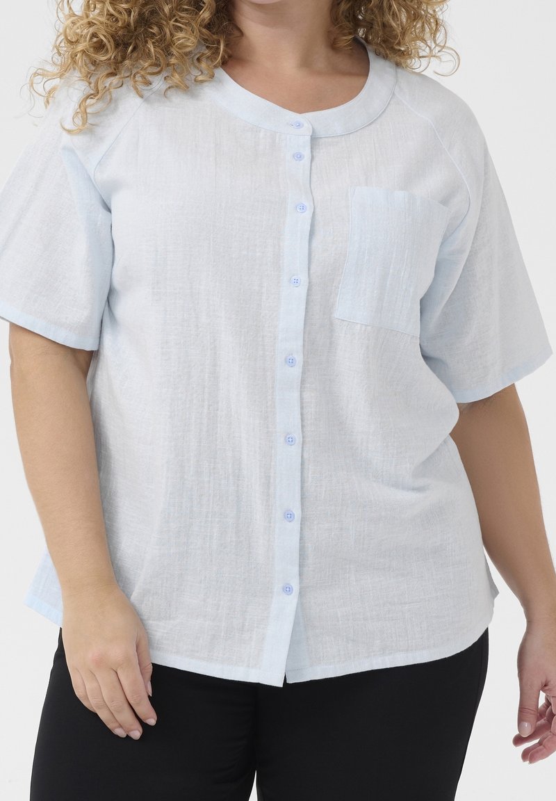 Personne aux cheveux bouclés portant une chemise bleu clair à manches courtes avec une poche poitrine et un pantalon noir sur un fond uni.