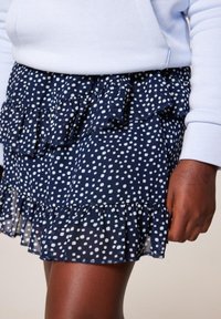 Námořnická modrá sukně s bílými puntíky, s volánkovými detaily a vrstveným designem. Vyrobena z lehké tkaniny s průhledným lemem.