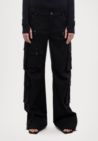 Svarta vida cargobyxor med flera fickor och dragkedjor, bärs tillsammans med svart topp och öppna skor som visar tårna.