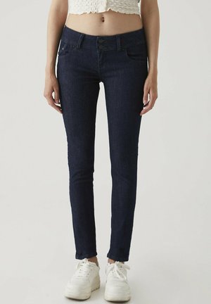 Dark blue skinny jeans with a high waist, two front pockets, and double button closure. Paired with white sneakers.