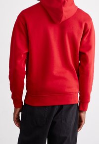 Felpa rossa con cappuccio regolabile, texture morbida, polsini e orlo a costine, indossata su pantaloni scuri. La vista da dietro mostra un tessuto liscio e privo di decorazioni.
