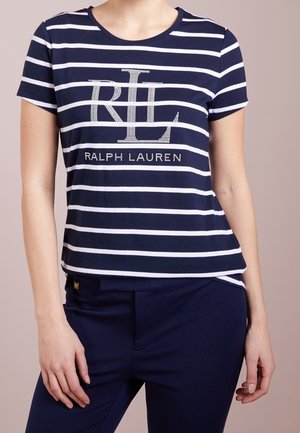 Femme portant un t-shirt Ralph Lauren rayé bleu marine et blanc et un pantalon bleu marine foncé, debout devant un fond uni.