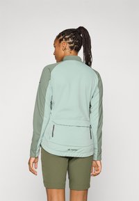 Person med flettehår iført en mintgrønn jakke og olivenfargede shorts, vendt bort, som viser jakkenes design og baklommen.