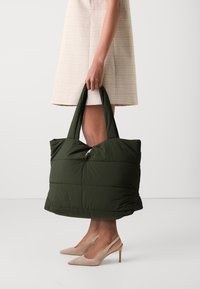 Sac fourre-tout vert rembourré avec fermeture éclair, doté de deux poignées robustes et d'une forme rectangulaire. Matériau en nylon texturé avec un design matelassé.