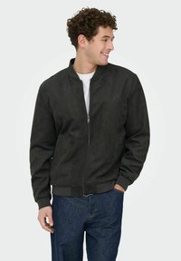 Homme souriant aux cheveux bouclés portant une veste noire zippée, une chemise blanche et un jean bleu, debout avec une main dans la poche, regardant vers la droite.