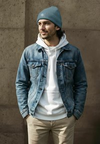 Blue denim jacket with chest pockets over a white hoodie, beige pants, and a light blue ribbed beanie with a compass logo.
