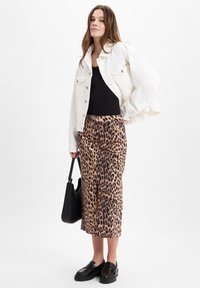 Leopardenmuster Midi-Rock mit einem Schlitz vorne, kombiniert mit einem schwarzen Rippenshirt und einer weißen Jeansjacke; accessoriert mit einer schwarzen Handtasche und klobigen Loafers.