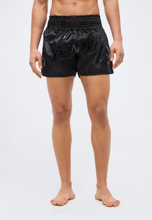 Męski tułów i nogi w czarnych błyszczących szortach z logo "VENUM" i wzorami błyskawic, stojący boso na jasnoszarym tle.