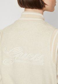 Gilet sans manches en polaire beige clair avec col et poignets côtelés. Présente un logo "Guess" brodé en blanc dans le dos, ajoutant du détail au design.