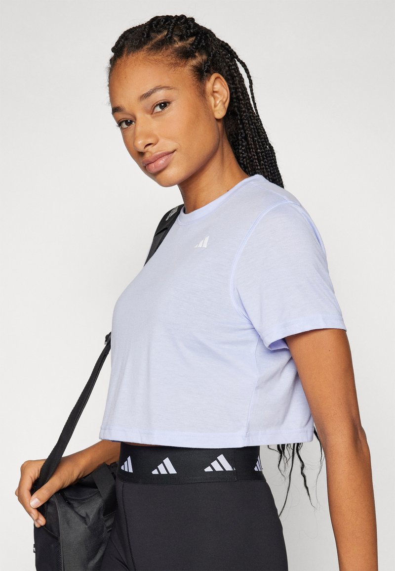 Camiseta corta de color púrpura claro con mangas cortas, textura suave, con un pequeño logo blanco en el pecho izquierdo, combinada con leggings negros.