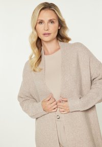 Béžový pletený kardigan s volným střihem, žebrovanými manžetami a otevřeným předním dílem, přehozený přes béžový top. Měkká textura, minimalistický design.