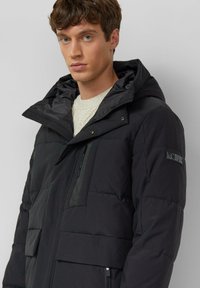 Schwarze Steppjacke mit Kapuze, großen Fronttaschen, einer Reißverschlusstasche an der Seite und einem Logoaufnäher am Ärmel. Glattes, strukturiertes Gewebe.