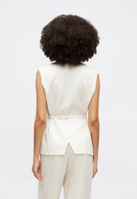 Top sans manches beige en tissu texturé, avec un nœud dans le dos et une fente au niveau de l'ourlet, associé à un pantalon de couleur claire.