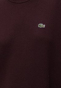 Tissu texturé bordeaux foncé avec un petit logo crocodile vert brodé sur la partie supérieure de la poitrine.