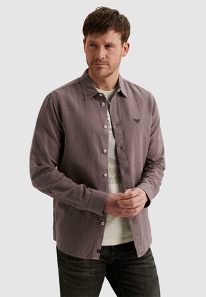 Muž s krátkymi hnedými vlasmi, oblečený v fialovastej košeli so zapnutými gombíkmi cez biele tričko s potlačou, stojaci so zloženými rukami.