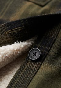 Veste à carreaux vert foncé et marron avec un bouton noir "Superdry" et une doublure en polaire blanche visible près de la boutonnière.