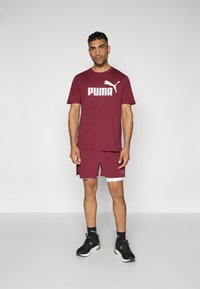 Bordeaux atletisk skjorte med hvid "PUMA" logo, parret med matchende shorts. Sorte sneakers og sorte sokker fuldender outfittet. Simpelt design.