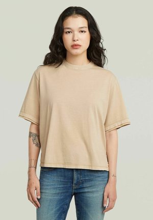 Giovane donna con lunghi capelli scuri che indossa una maglietta beige oversize e jeans blu, in piedi con le braccia rilassate lungo i fianchi.