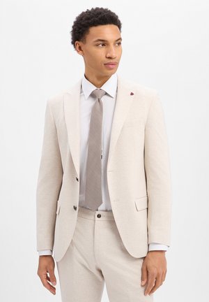 Man wearing a light beige suit with a white shirt and textured beige tie, standing and looking slightly to his right.