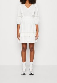 Mujer vistiendo un vestido blanco a la altura de la rodilla con detalles de encaje, mangas abullonadas y volantes, combinado con calcetines blancos hasta el tobillo y zapatillas deportivas.