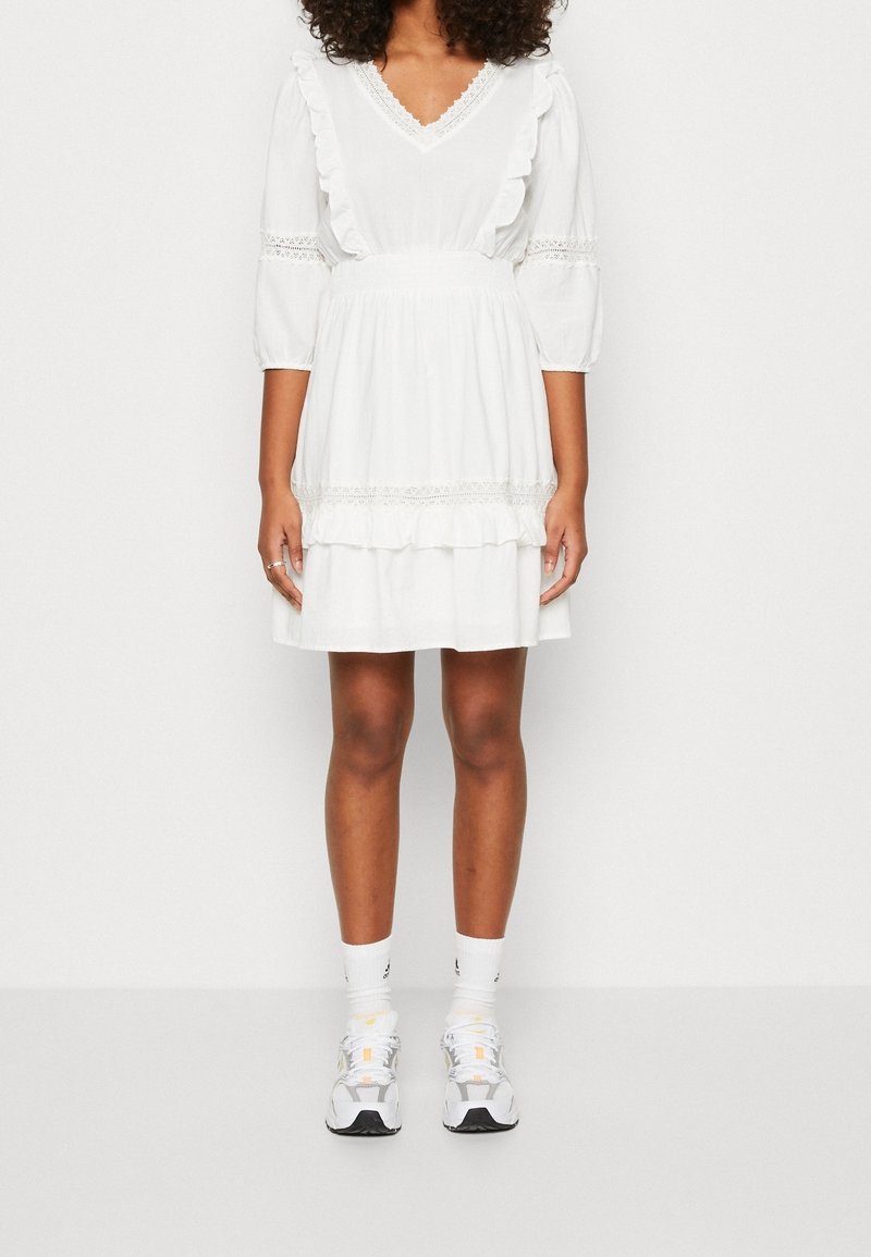 Mujer vistiendo un vestido blanco a la altura de la rodilla con detalles de encaje, mangas abullonadas y volantes, combinado con calcetines blancos hasta el tobillo y zapatillas deportivas.