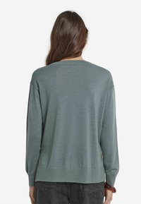 Kvinna med långt brunt hår som bär en löst sittande grön tröja och mörkgrå jeans, stående med ryggen mot en enkel bakgrund.