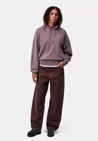 Sudadera de lavanda oscuro con bolsillo frontal y capucha con cordón, combinada con pantalones marrones sueltos y zapatos negros, de pie contra un fondo neutro.