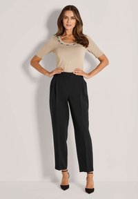 Vrouw met lang haar in een beige top met korte mouwen en versieringen, gecombineerd met hoge zwart broek, staand met handen op de heupen.