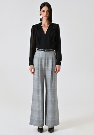 Blusa nera con dettagli arricciati, abbinata a pantaloni grigi a quadri con gamba larga, caratterizzati da una cintura nera e scarpe con tacco quadrato.