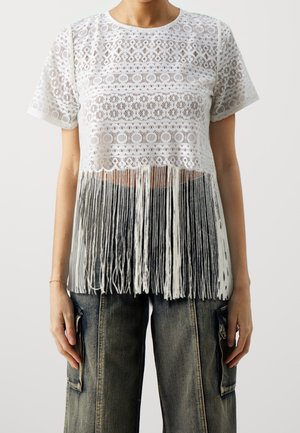 Kvinde iført en hvid blonde-croptop med frynsekant kombineret med løse, falmede sorte cargobukser, stående mod en ensfarvet baggrund.