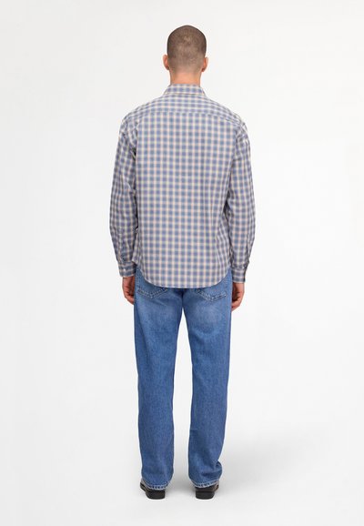 Homme debout, vu de dos, portant une chemise à manches longues à carreaux bleu et beige, un jean en denim bleu et des chaussures noires.