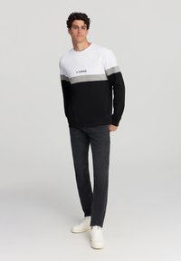 Uomo in piedi che indossa un sweatshirt a blocchi di colore con la scritta "5th Avenue", jeans neri e sneakers bianche, su uno sfondo chiaro e uniforme.