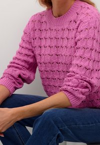 Rosa gestrickter Pullover mit einem strukturierten, offenen Muster und gerippten Bündchen. Kombiniert mit blauen Jeans für einen Kontrast.