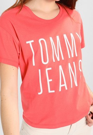 Camiseta de algodón color coral con un gran logo blanco de "TOMMY JEANS". El diseño incluye mangas cortas y un cuello redondo.