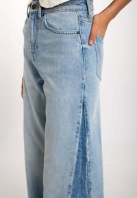 Vaqueros acampanados de color azul claro con una abertura lateral, que presentan un diseño clásico de cinco bolsillos y un sutil desvanecimiento en la tela.