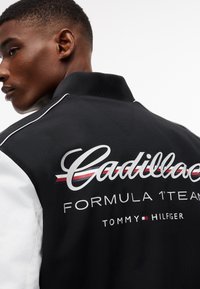 Mladý muž s krátkymi vlasmi, oblečený v čierno-bielej bunde s logom "Cadillac Formula 1 Team Tommy Hilfiger" na chrbte.
