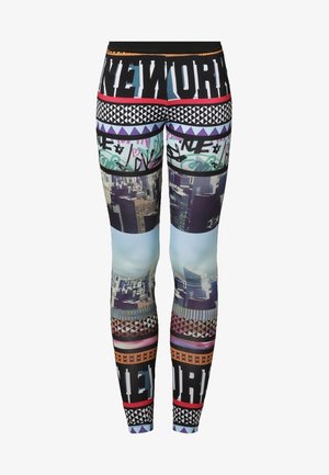 Vrouw draagt een donker denimjack, zwarte top, kleurrijke legging met stadsgezichten en witte veterschoenen, staand op een lichte achtergrond.