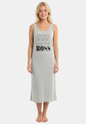Frau steht barfuß, trägt ein ärmelloses graues Kleid mit dem Text "ICH BIN NICHT BOSSY, ICH BIN DER BOSS" auf der Vorderseite.